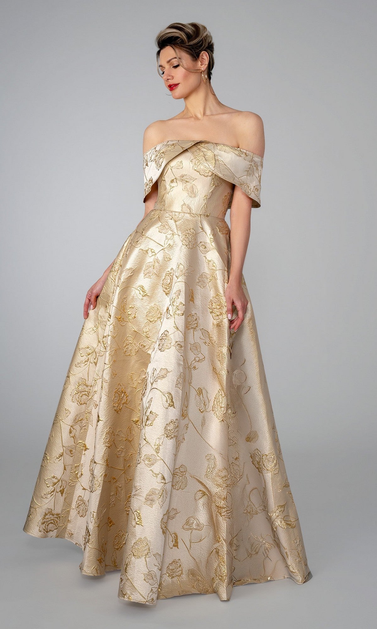 A woman in the Stella Couture 26313 long formal dress, featuring a champagne gold floral brocade and off-the-shoulder design, stands against a plain light gray background. The floor-length gown exudes elegant, timeless sophistication.